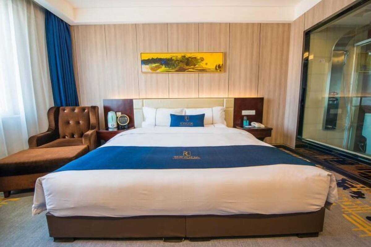 Une chambre d’hôtel avec un grand lit, une chaise, une table de chevet, un tableau au mur et une cloison en verre.