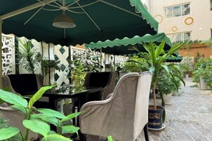Un área de estar al aire libre con mesas y sillas, rodeada de plantas en macetas y una pared decorativa.
