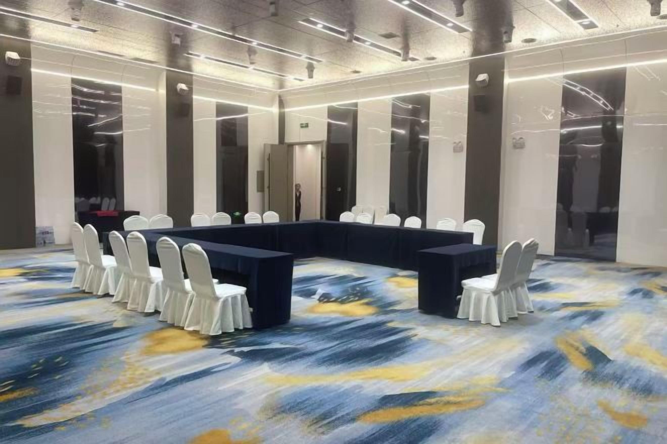 A conference room with a U-shaped table, white chairs, and a patterned carpet.