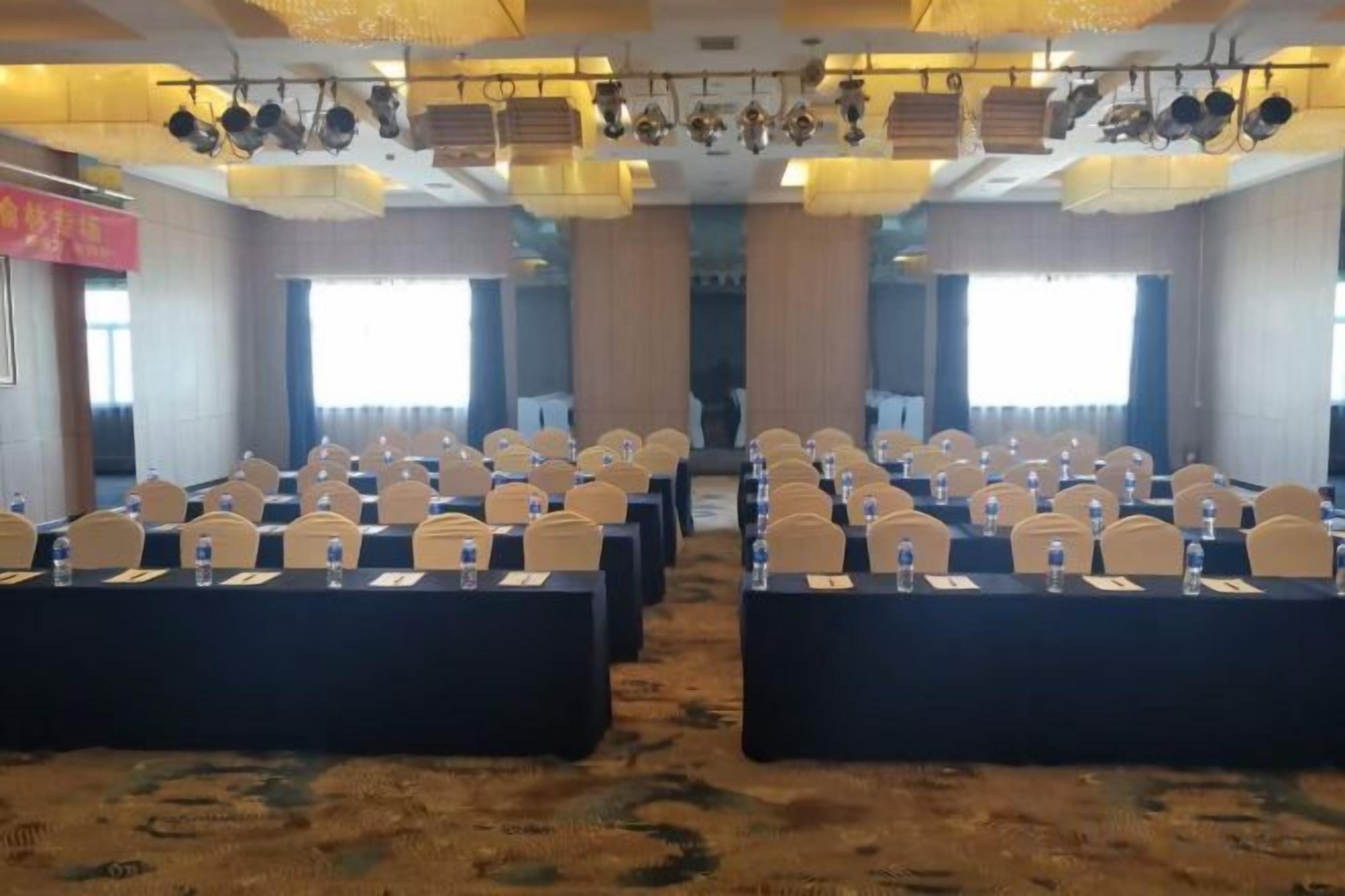 Une salle de conférence avec des rangées de tables et de chaises, de grandes fenêtres et un sol recouvert de moquette.