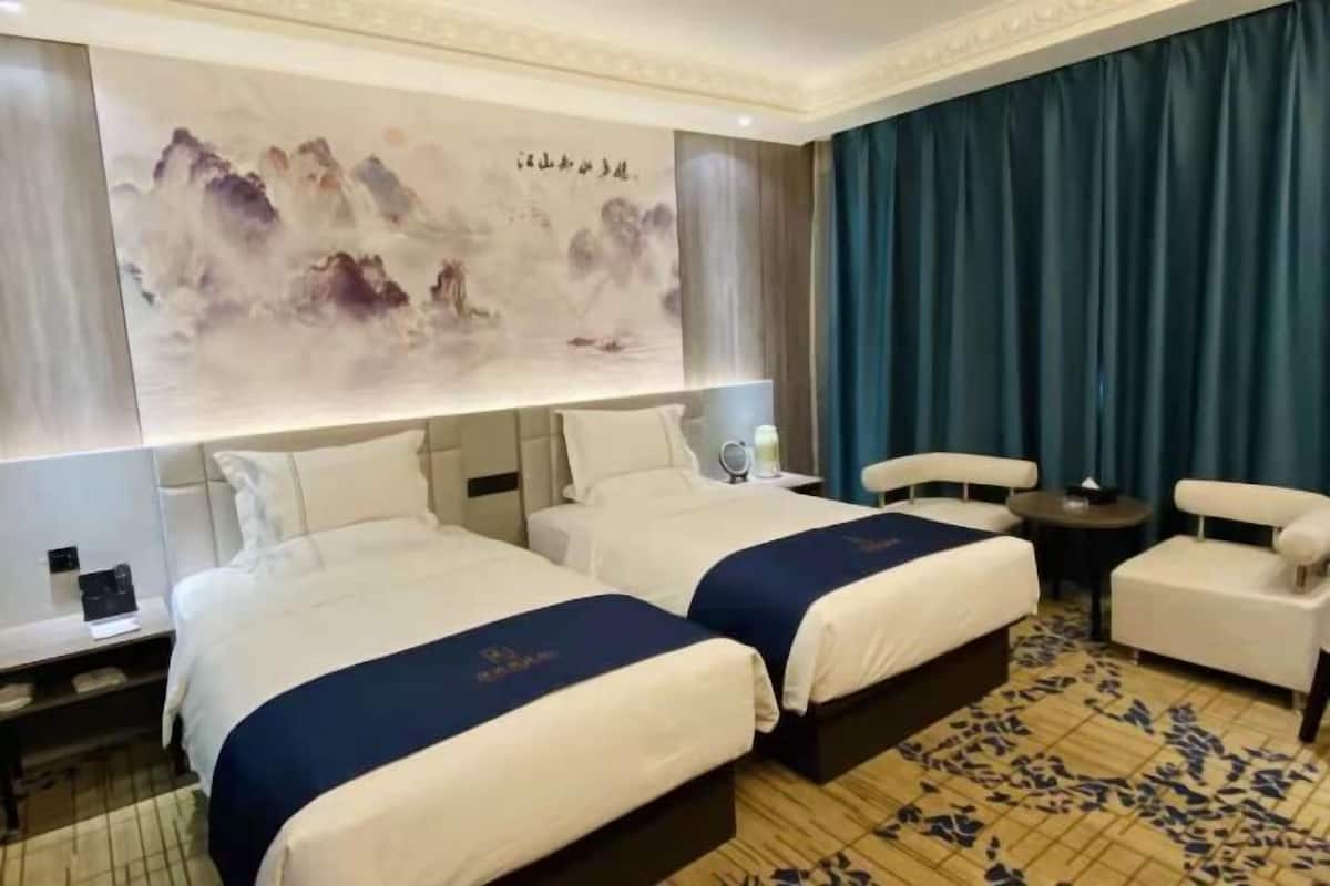 Une chambre d’hôtel avec deux lits, une grande peinture murale, une table ronde et un rideau bleu.