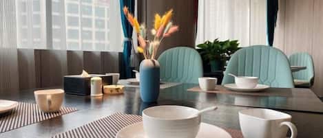 A dining area with a table set for two, a vase with dried flowers, and a city view through the window.