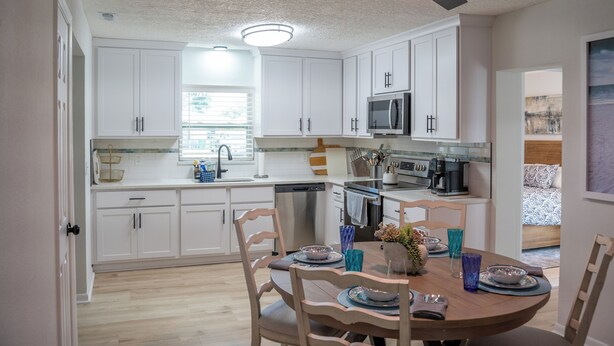 Una cocina moderna con armarios blancos, electrodomésticos de acero inoxidable y un comedor con mesa y sillas de madera.