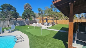 Un jardin avec une piscine, une pelouse artificielle et un espace patio aménagé avec du mobilier d’extérieur.