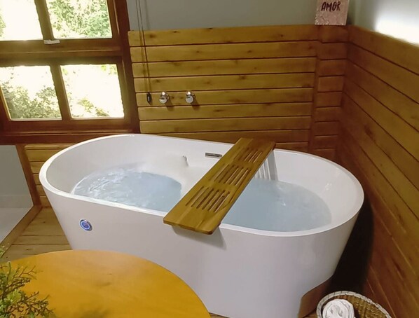 Una cabaña de madera con una bañera grande, un banco de madera y una ventana con vistas a la vegetación.