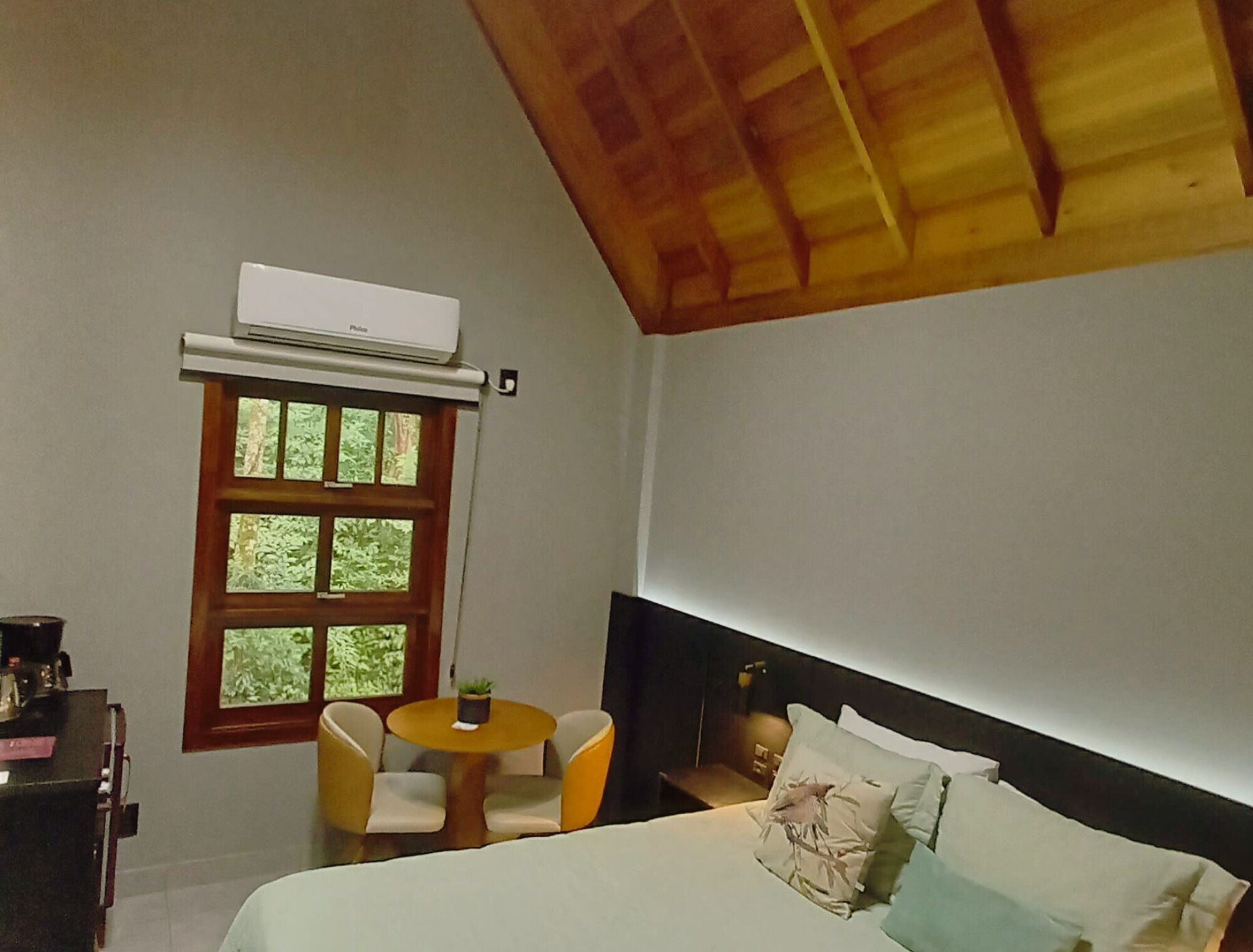 Un dormitorio con cama, una mesita redonda con sillas, una ventana con vista a árboles y un equipo de aire acondicionado.