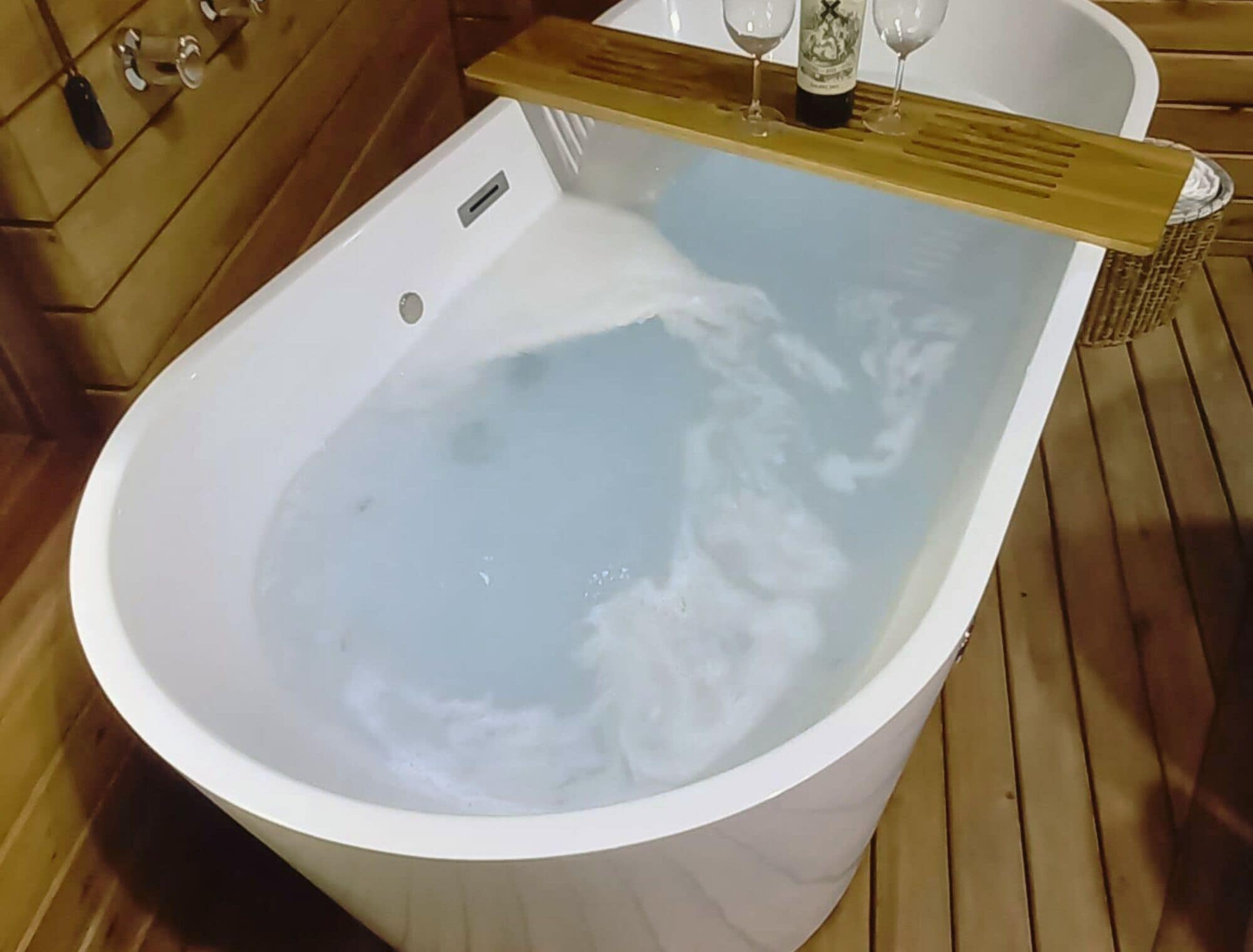 Una bañera llena de agua, suelo de madera y una botella de vino sobre una estantería de madera.