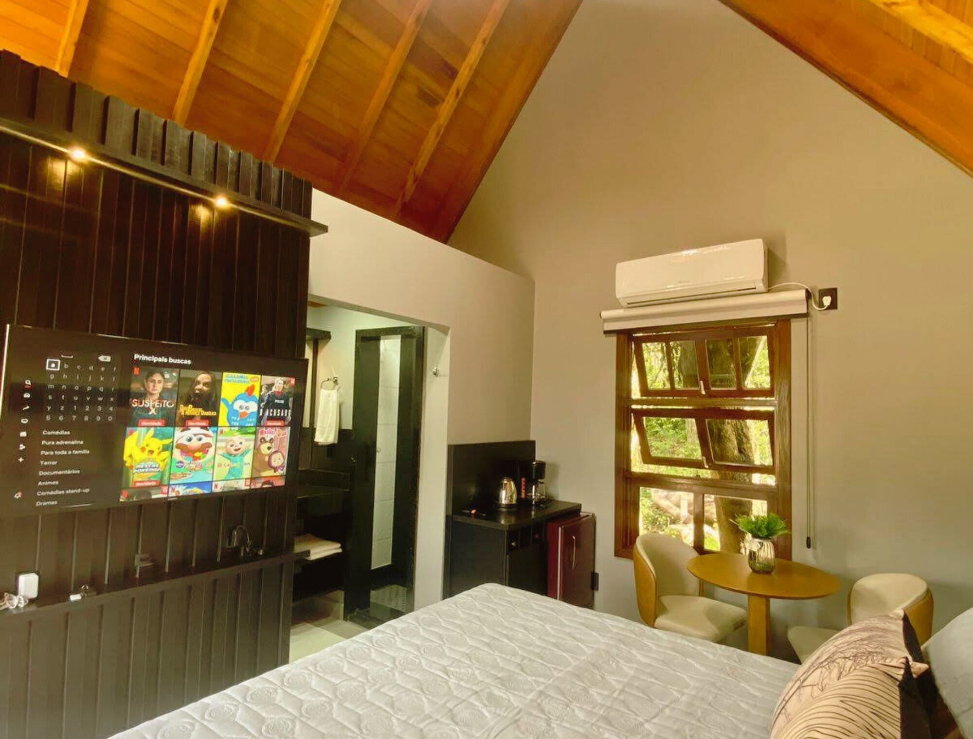 A hotel room with a bed, a small table with chairs, a TV mounted on the wall, and a window with a view of greenery.