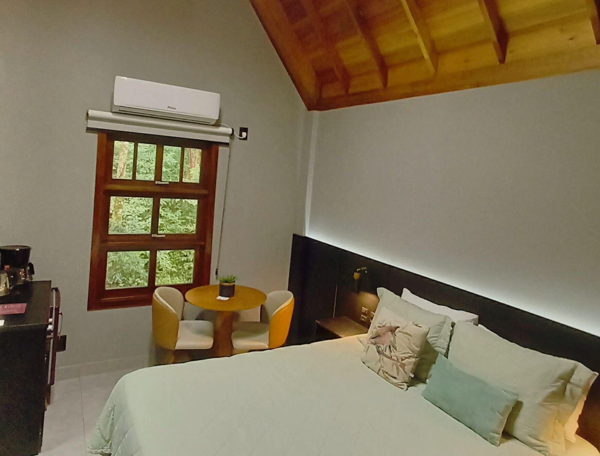 A bedroom with a bed, a small round table with chairs, a window with a view of greenery, and an air conditioning unit.