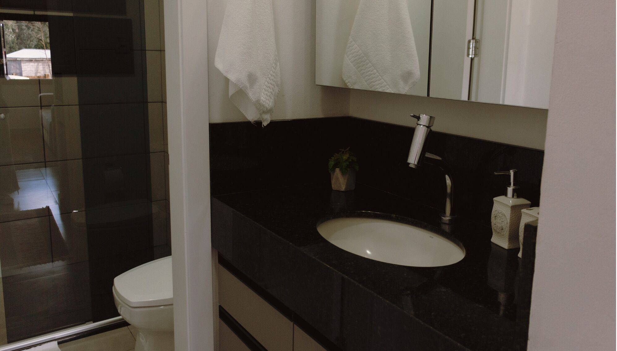 Un baño moderno con encimera negra, lavabo blanco y un inodoro. Hay una planta pequeña en la encimera y una toalla colgada en la pared.