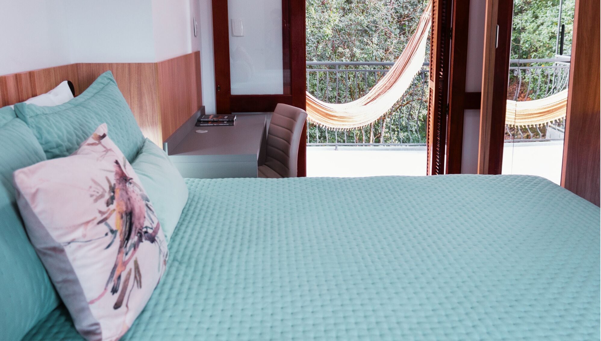 Un dormitorio con cama, almohadas, un escritorio y una hamaca fuera de la ventana.