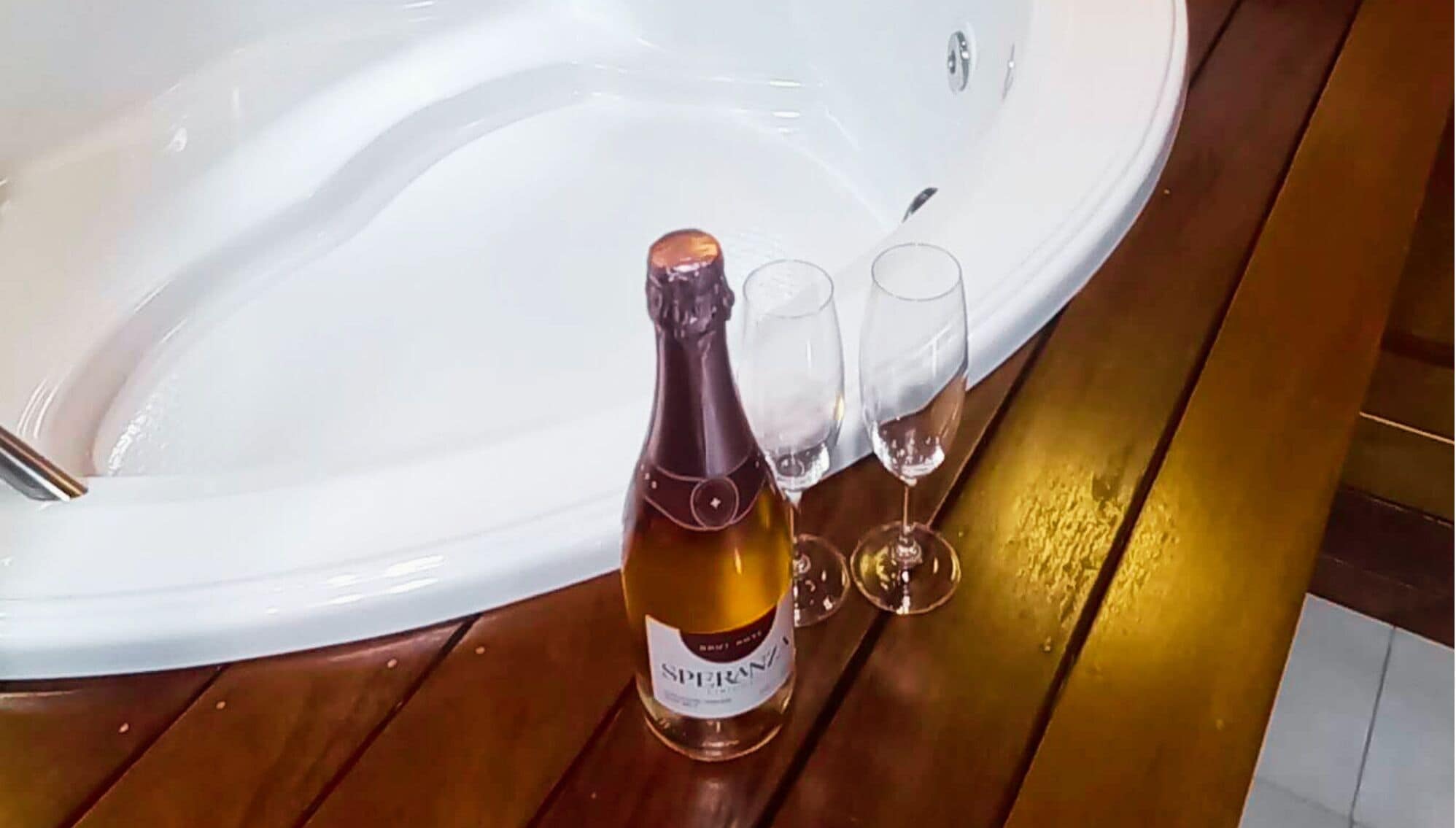 Une bouteille de champagne et deux verres sur une surface en bois, près d’une baignoire blanche.