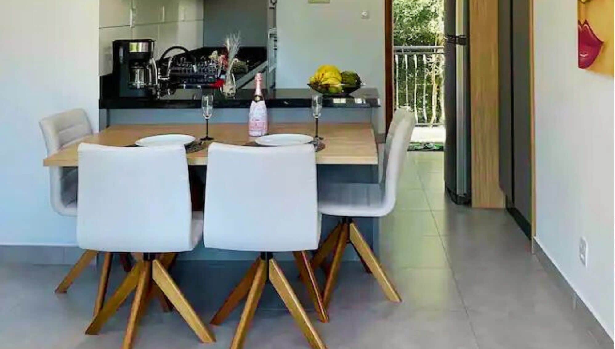 Un comedor con una mesa y sillas de madera, una cocina al fondo y vistas al exterior a través de un ventanal.