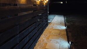A wooden fence with string lights and stepping stones leading to a dark area.