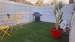 A backyard with artificial grass, a barbecue grill, yellow chairs, and a red potted plant. - Small Love/BBQ (Lehigh Acres)