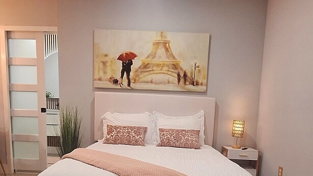A bedroom with a bed, a nightstand, a lamp, a painting of the Eiffel Tower, and a door with frosted glass panels. - Small Love/BBQ (Lehigh Acres)