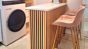 A modern kitchen with a built-in washing machine, a wooden island with bar stools, and a shelf unit with decorative items. - Small Love/BBQ (Lehigh Acres)