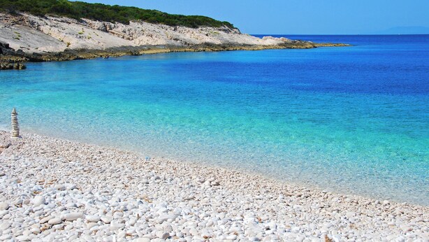 Une plage de galets bordée d’une eau turquoise cristalline, avec une falaise rocheuse en arrière-plan.