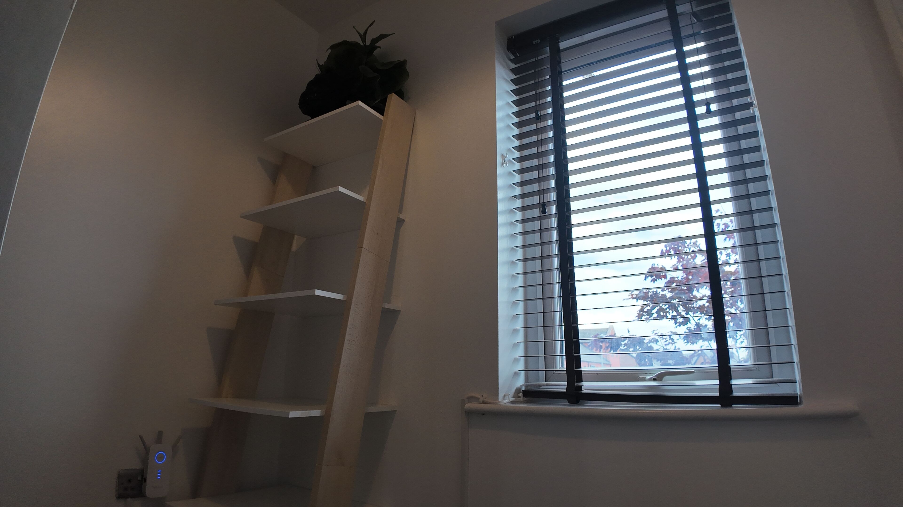 Een kamer met een raam met jaloezieën, een witte plankkast en een wandcontactdoos.