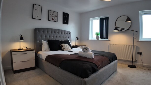 A modern bedroom with a tufted headboard, a bedside table with a lamp, a large window with curtains, and artwork on the wall. - Stylish Stay in Vibrant Salford, Manchester! (Salford)