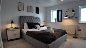 A modern bedroom with a tufted headboard, a bedside table with a lamp, a large window with curtains, and artwork on the wall. - Stylish Stay in Vibrant Salford, Manchester! (Salford)