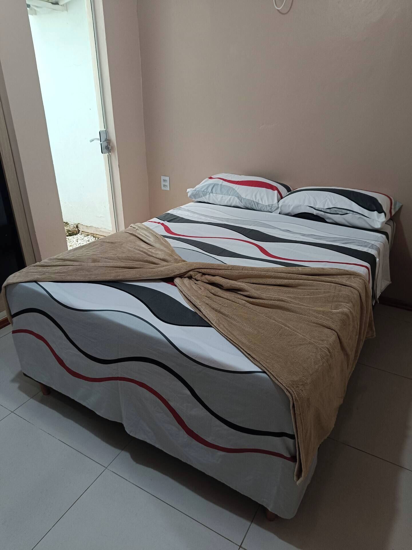 A bed with a patterned bedspread and a brown blanket, a white wall, and a door leading outside.