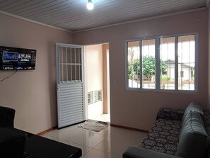 Una sala de estar con sofá, televisor montado en la pared, puerta con ventanal y suelo de baldosas.