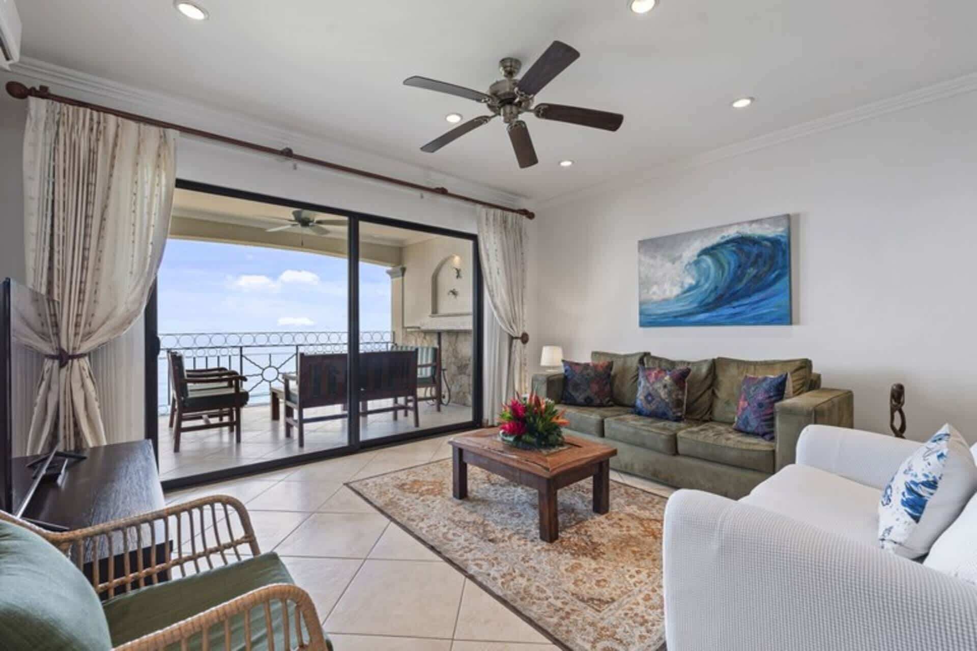 A spacious living area with a balcony, a ceiling fan, a sofa set, and a painting of a wave.