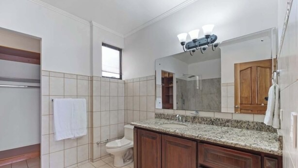 A bathroom with a toilet, a sink with a granite countertop, wooden cabinets, and a large mirror above the sink. - Stunning Condo with Panoramic Beach, Ocean, and Mountain Views in Playa Flamingo (Playa Flamingo)
