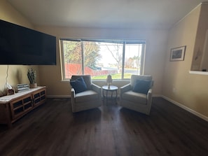 A living room with two armchairs, a small table, a TV mounted on the wall, and a window with a view of a backyard.