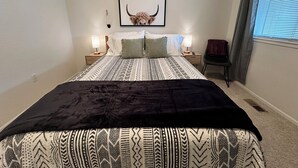 Un dormitorio con una colcha estampada, un cuadro en la pared, lámparas de noche, una silla y una ventana con persianas.
