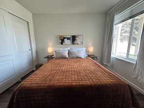 A bedroom with a large bed, two bedside tables with lamps, a door, a window, and a painting on the wall.