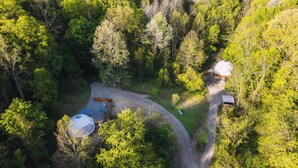 Une vue aérienne d’une zone boisée où l’on aperçoit deux structures en forme de dôme, vraisemblablement des yourtes, reliées par un sentier sinueux.