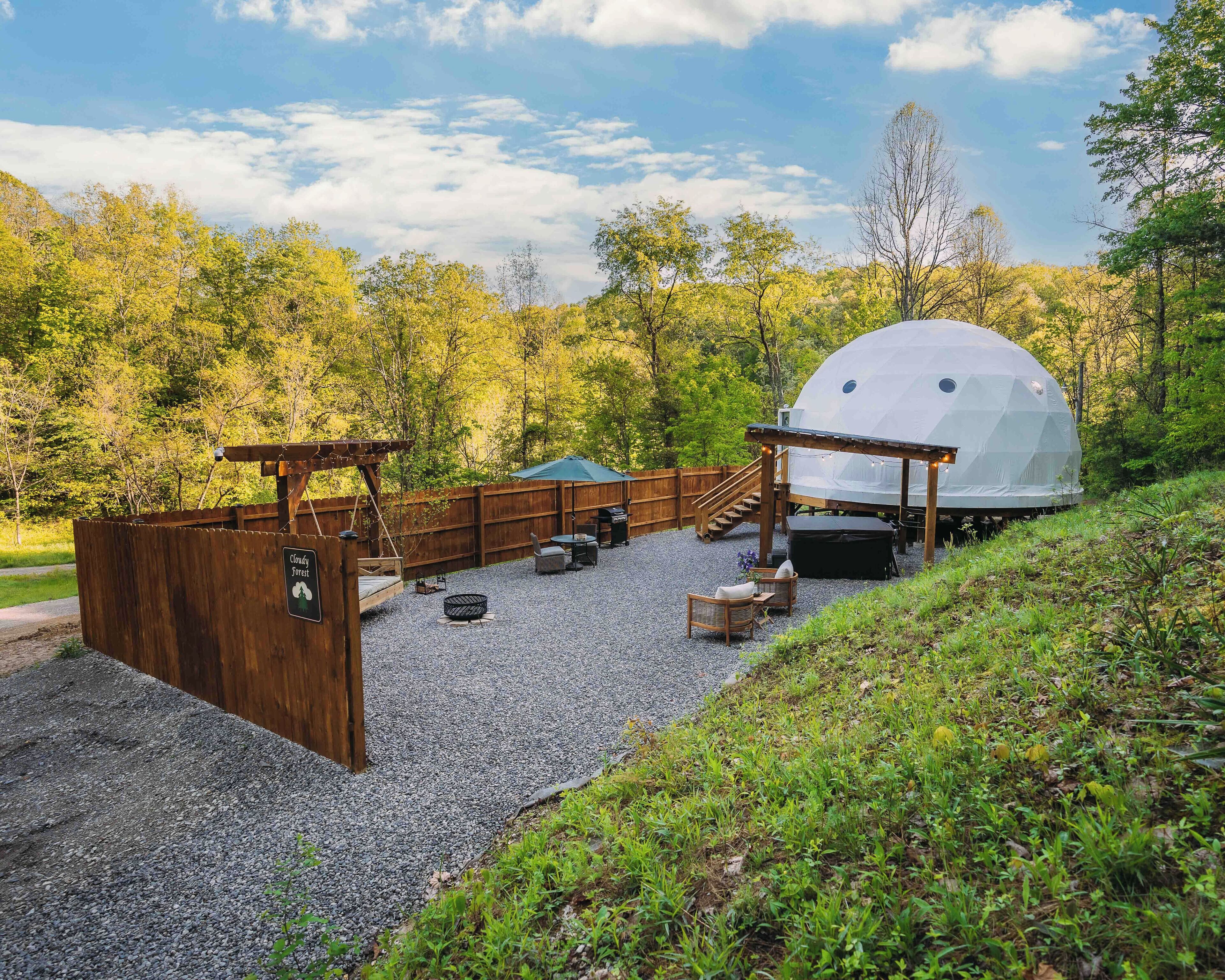 A geodesic dome structure in a forested area with a wooden deck, outdoor seating, and a gravel path.