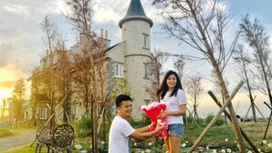 A man kneeling and presenting a red gift to a woman in front of a castle-like building with a tower.