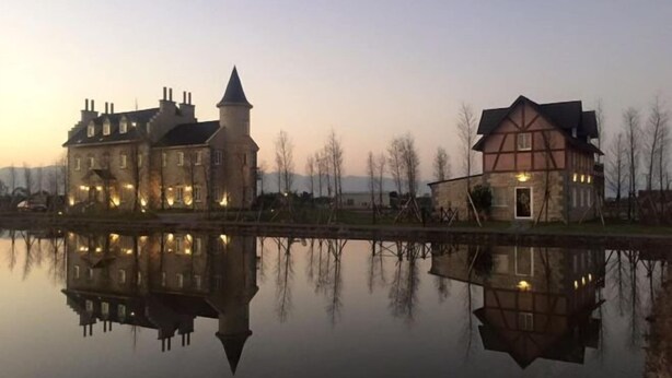 A large, historic building with a tower and a smaller adjacent structure, both reflecting on a calm body of water.