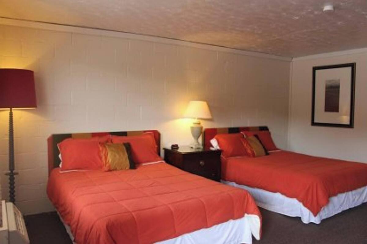 A hotel room with two beds, each with red bedding and white sheets. There is a nightstand with a lamp between the beds.