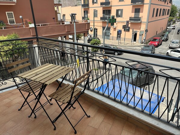 A balcony with a wooden table and chairs, a metal railing, and a view of a street with parked cars and buildings. - Room25 InCentro Guest House (Cercola)