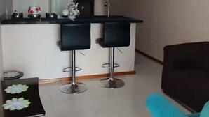 Una sala de estar moderna con un sofá azul, una mesita con objetos decorativos, y una cocina con un taburete de bar y un refrigerador.
