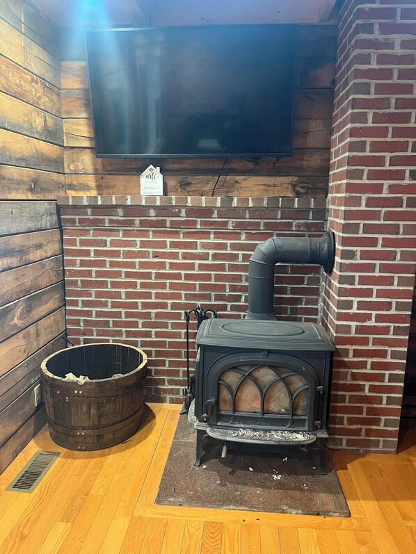 A wood-burning stove with a chimney, a wooden barrel, and a television mounted on a brick wall.