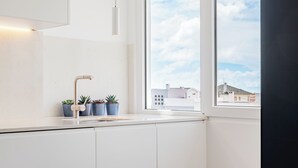 Una cocina moderna con armarios blancos, encimera blanca, una ventana con vistas a edificios y tres macetas con plantas.