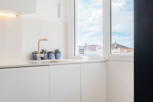 Una cocina moderna con armarios blancos, encimera blanca, una ventana con vistas a edificios y tres macetas con plantas.