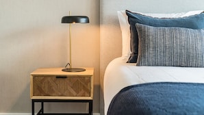 Una cama bien hecha con edredón blanco y almohadas a rayas azules, una mesita de noche de madera pequeña con una lámpara negra, y una pared lisa al fondo.
