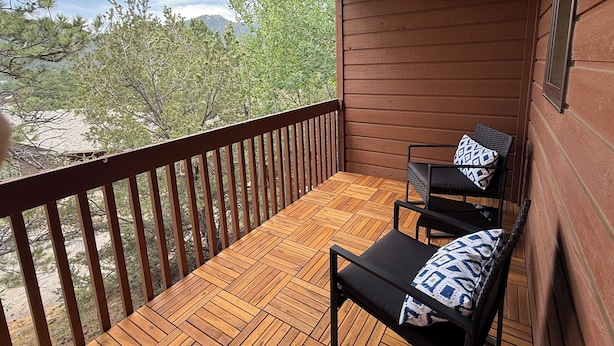 Eine Holzterrasse mit Geländer, zwei schwarze Stühle mit gemusterten Kissen und Blick auf Bäume und Berge.