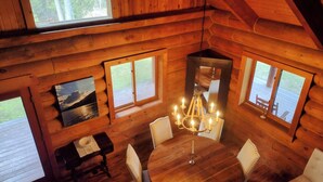 A cozy cabin with a wooden interior, a round dining table, and multiple windows offering views of the surrounding forest.