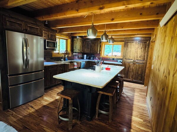 A spacious kitchen with wooden cabinets, a stainless steel refrigerator, a large island with stools, and pendant lights.