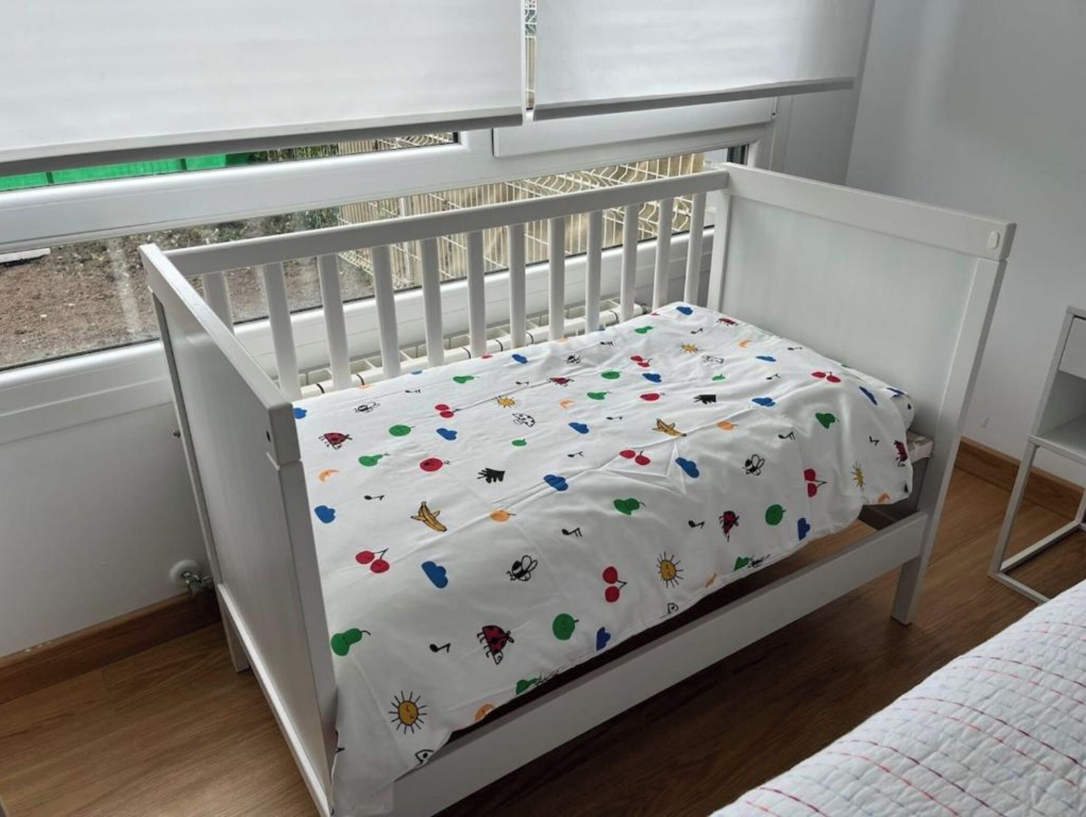 A white crib with a colorful mattress, positioned near a window with a view of a building outside.