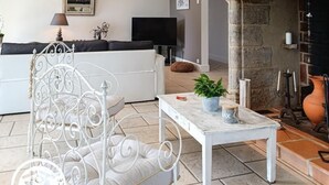 A living room with a white sofa, a white metal chair, a small table with a plant, and a stone fireplace. - Gîtes de France® - Lyna's Villa (PUYBRUN)