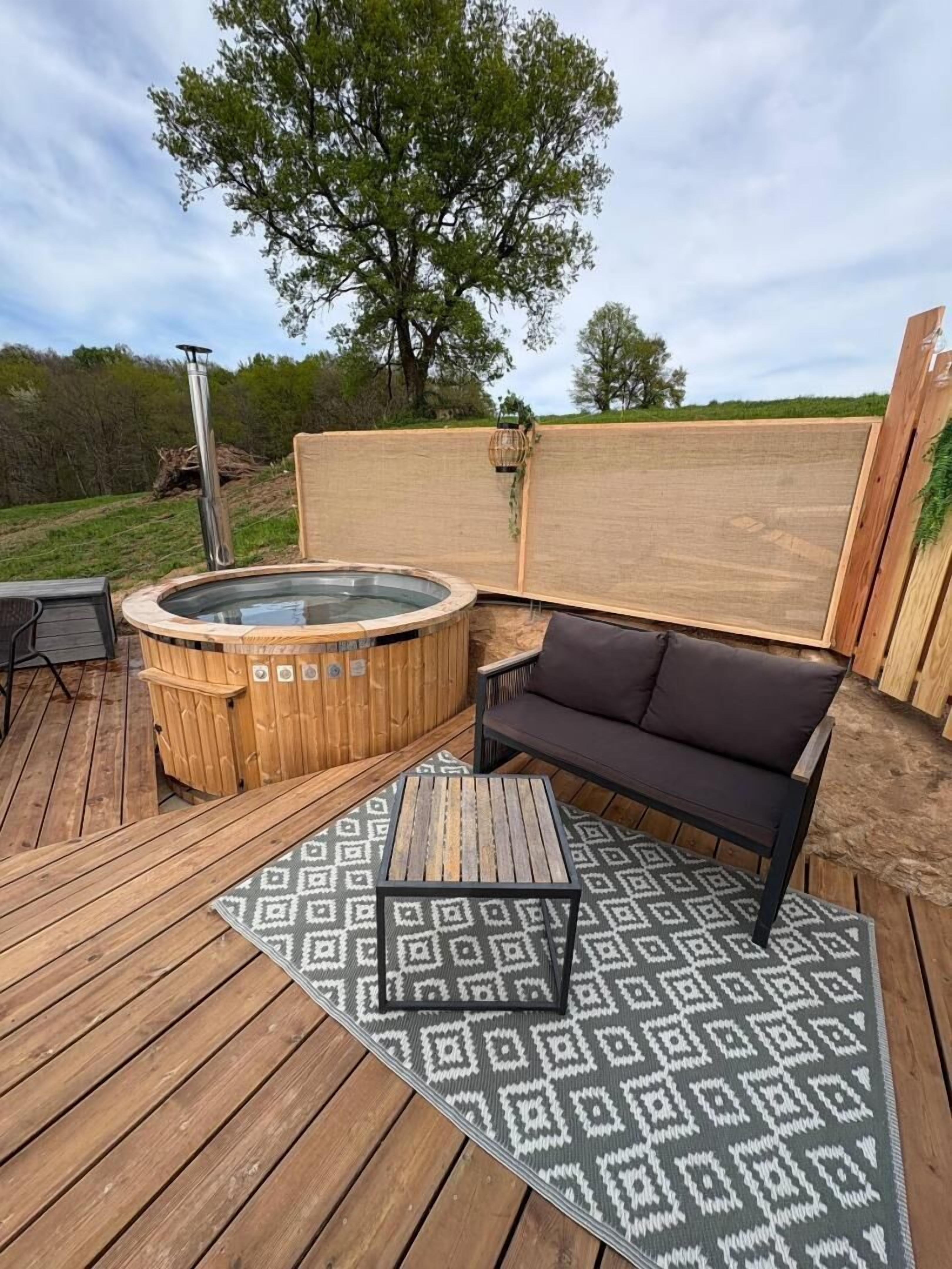 Un terrazzo in legno con una vasca idromassaggio, un divano nero e un tappeto decorato.