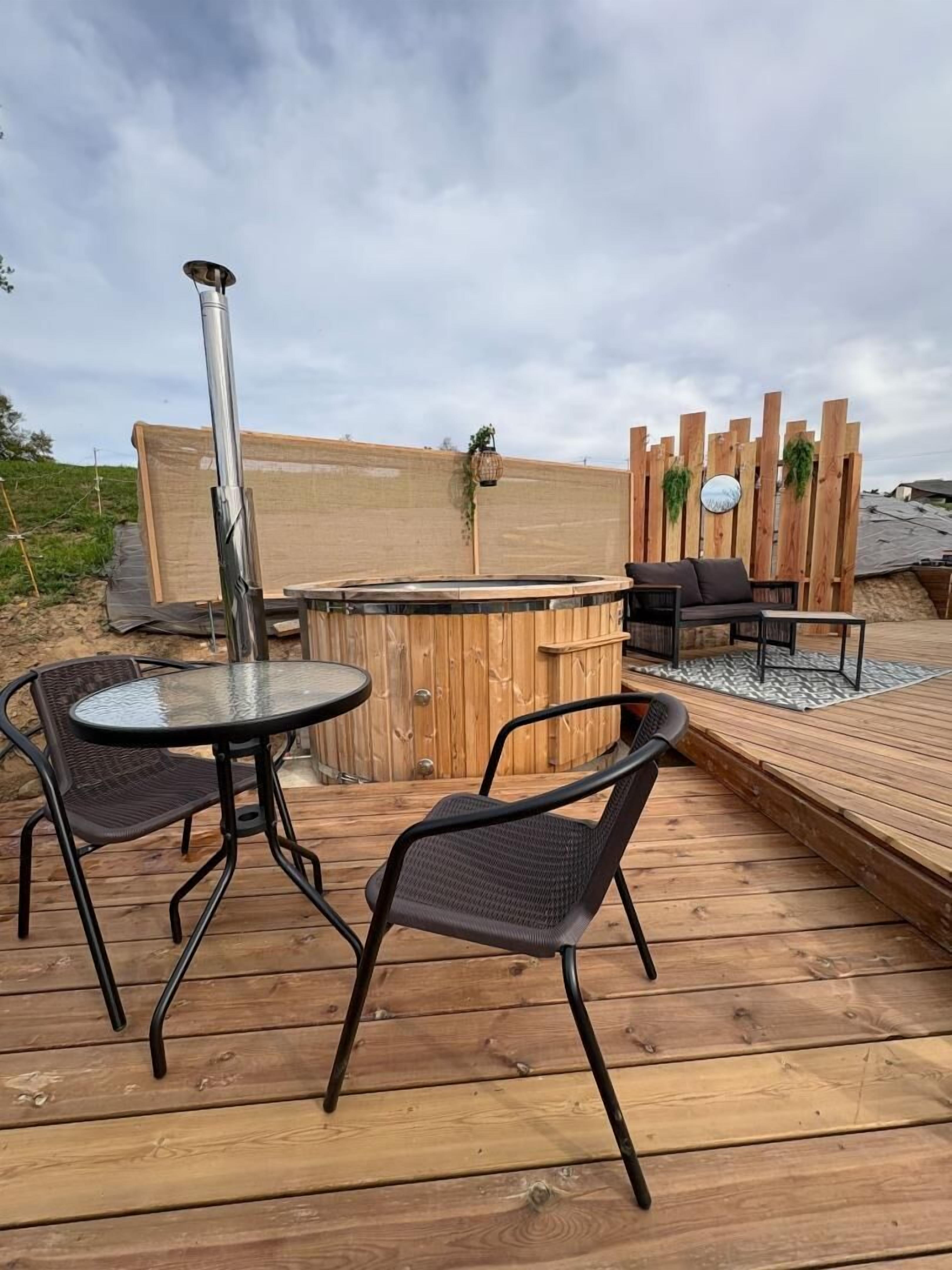 A wooden deck with a glass-top table and two chairs, a hot tub, and a wooden fence with hanging plants.